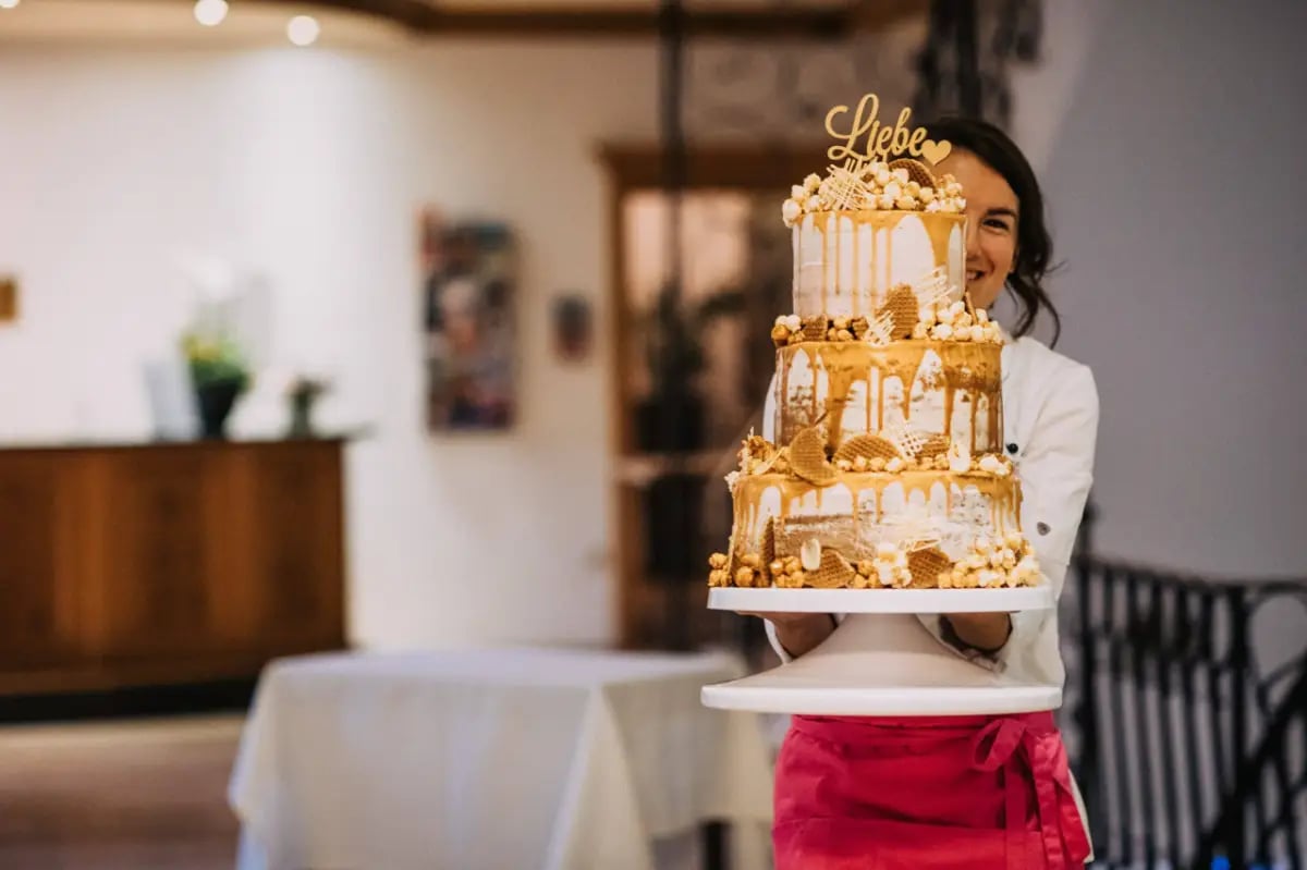 Eine Person hält eine große, dreistöckige Torte mit goldenen und weißen Dekorationen auf einem weißen Podest.