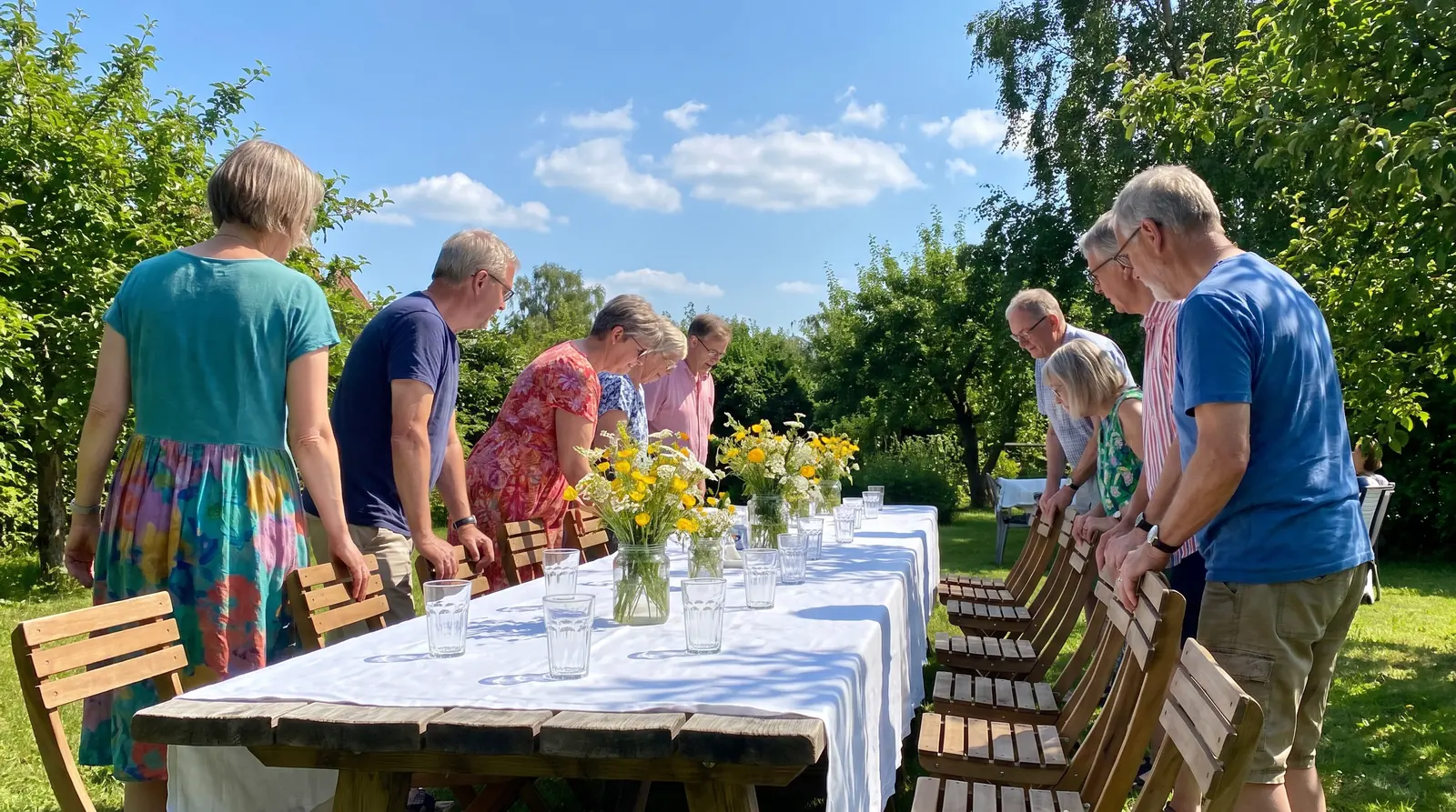 Gäste stellen im sonnigen Garten Stühle an eine lange Festtafel mit Wildblumen, so kann ein geplantes Event aussehen