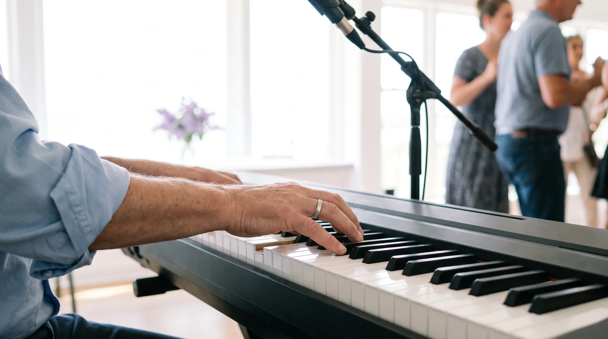 Hand eines Musikers mit Ehering auf den Tasten eines schwarzen Keyboards, dahinter ein Mikrofon am Ständer und tanzende Gäste im hellen Saal
