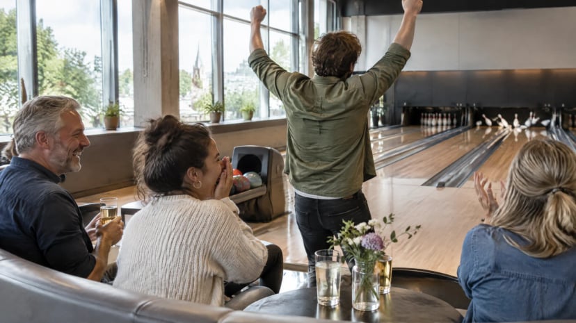 Gruppenaktivitäten beim Bowling: Freunde auf der Sitzbank jubeln über einen Strike, im Hintergrund die Bahnen und große Fenster mit Tageslicht