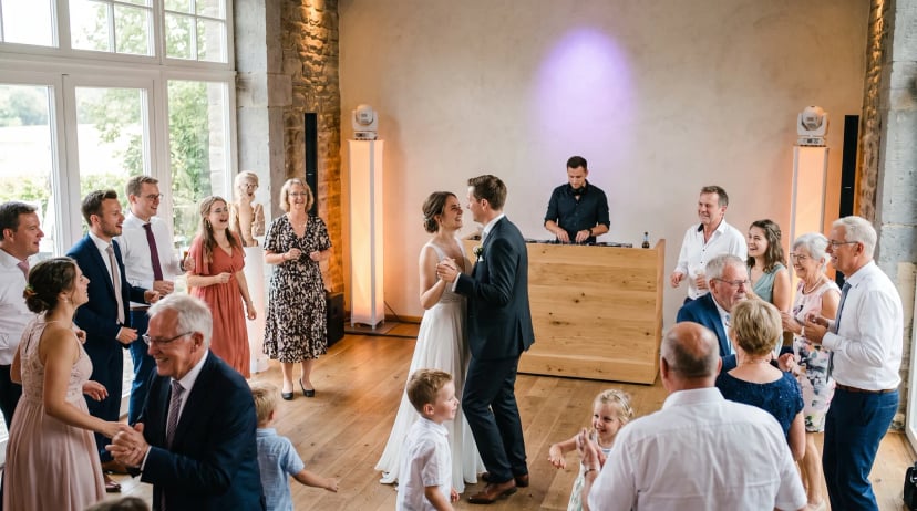 Hochzeits-DJ Rheinbach: DJ mit Kopfhörern hinter modernem Pult, Gäste in Festkleidung tanzen und lachen auf der Tanzfläche in warmem Licht.