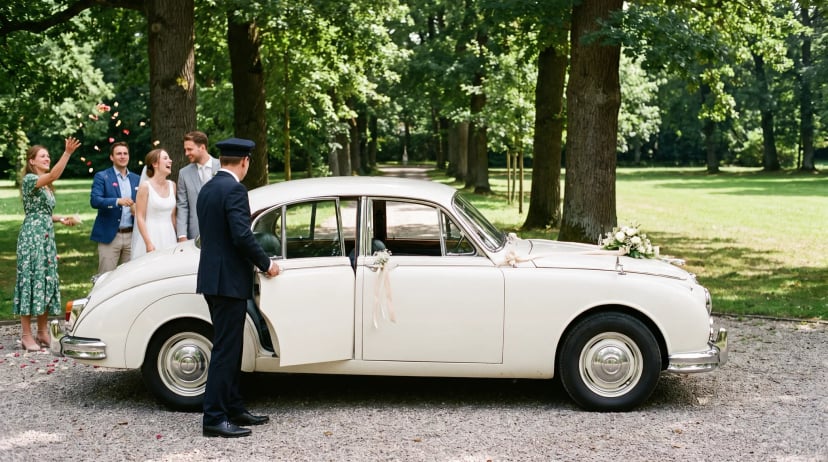 Hochzeitsauto mieten: cremefarbener Oldtimer mit Blumenschmuck in der Parkallee, der Chauffeur hält dem Brautpaar die Tür auf