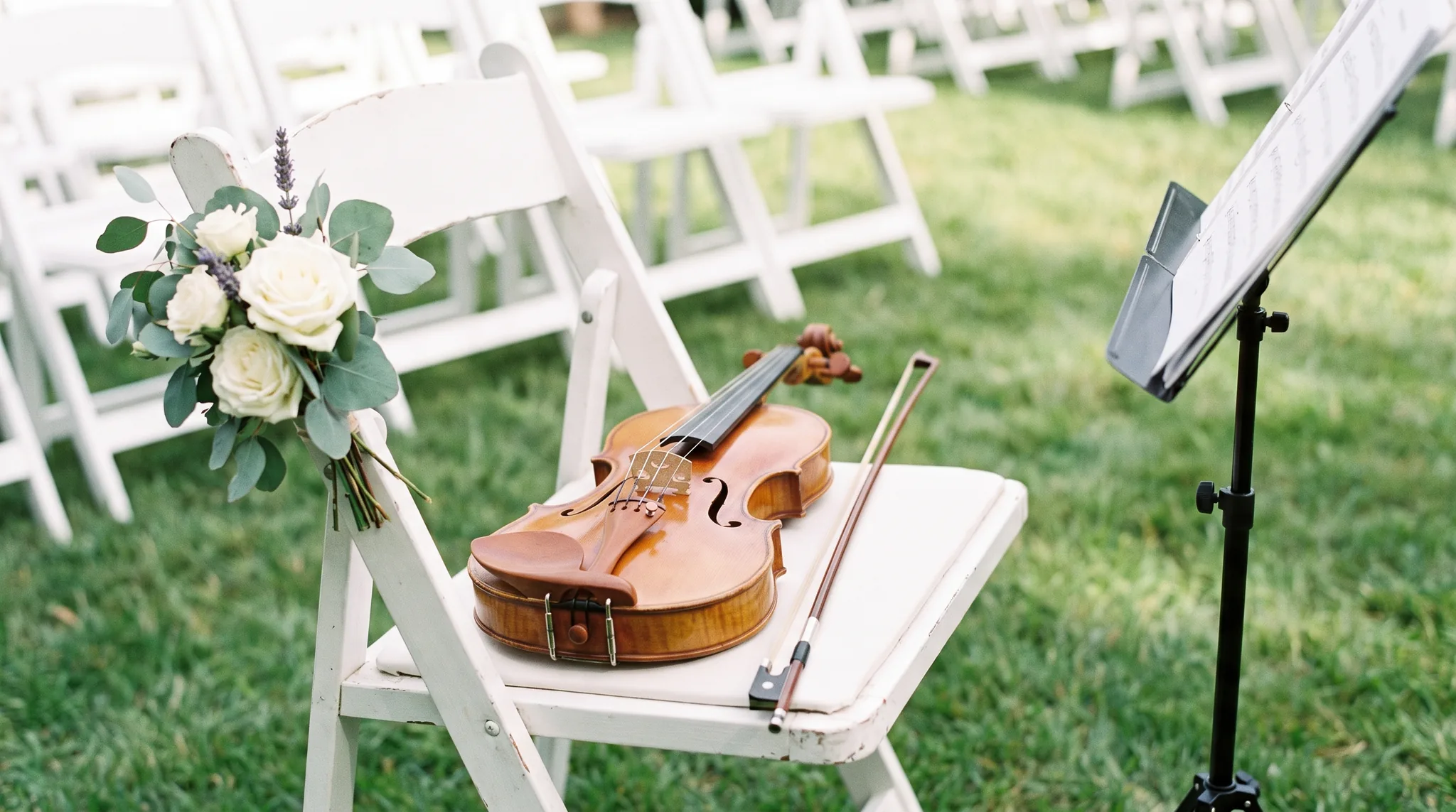 Hochzeitsmusiker Detail: Geige mit Bogen auf einem weißen Stuhl der Trauung im Garten neben Notenständer und Strauß aus weißen Rosen mit Lavendel