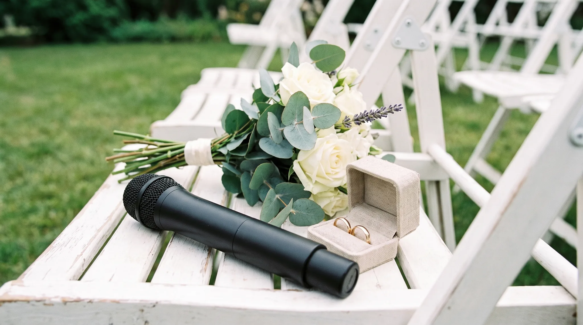 Hochzeitssängerin Detail: schwarzes Mikrofon auf einem weißen Stuhl der freien Trauung neben Brautstrauß aus weißen Rosen und Eheringen