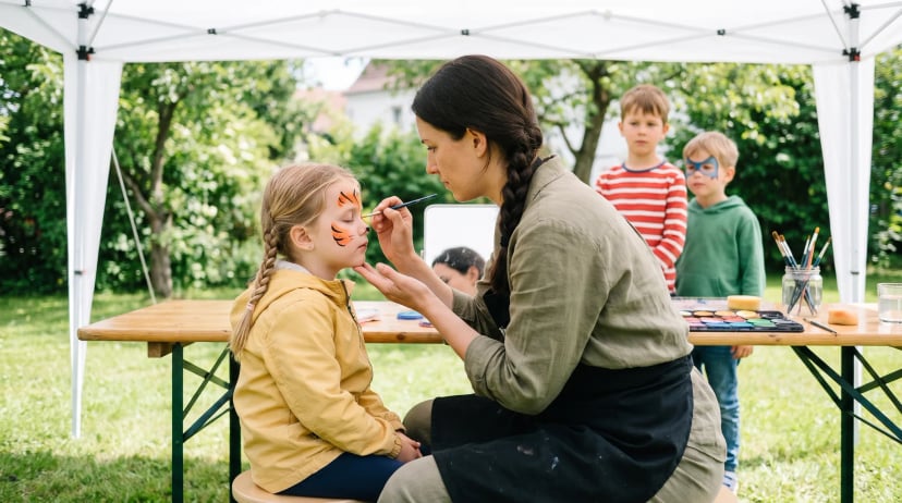 Kinderschminken buchen: Eine Schminkerin malt einem Mädchen mit geschlossenen Augen Tigerstreifen ins Gesicht, unter einem Pavillon im Garten, zwei Jungen warten im Hintergrund
