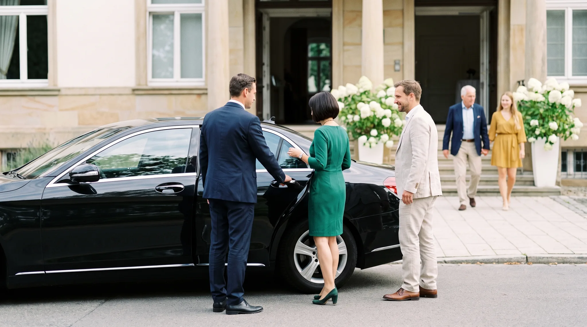 Limousinenservice vor einer Villa: Chauffeur im dunklen Anzug öffnet einer Frau im grünen Kleid die Tür einer schwarzen Limousine, ihr Begleiter wartet daneben, im Hintergrund kommen Gäste die Treppe herunter