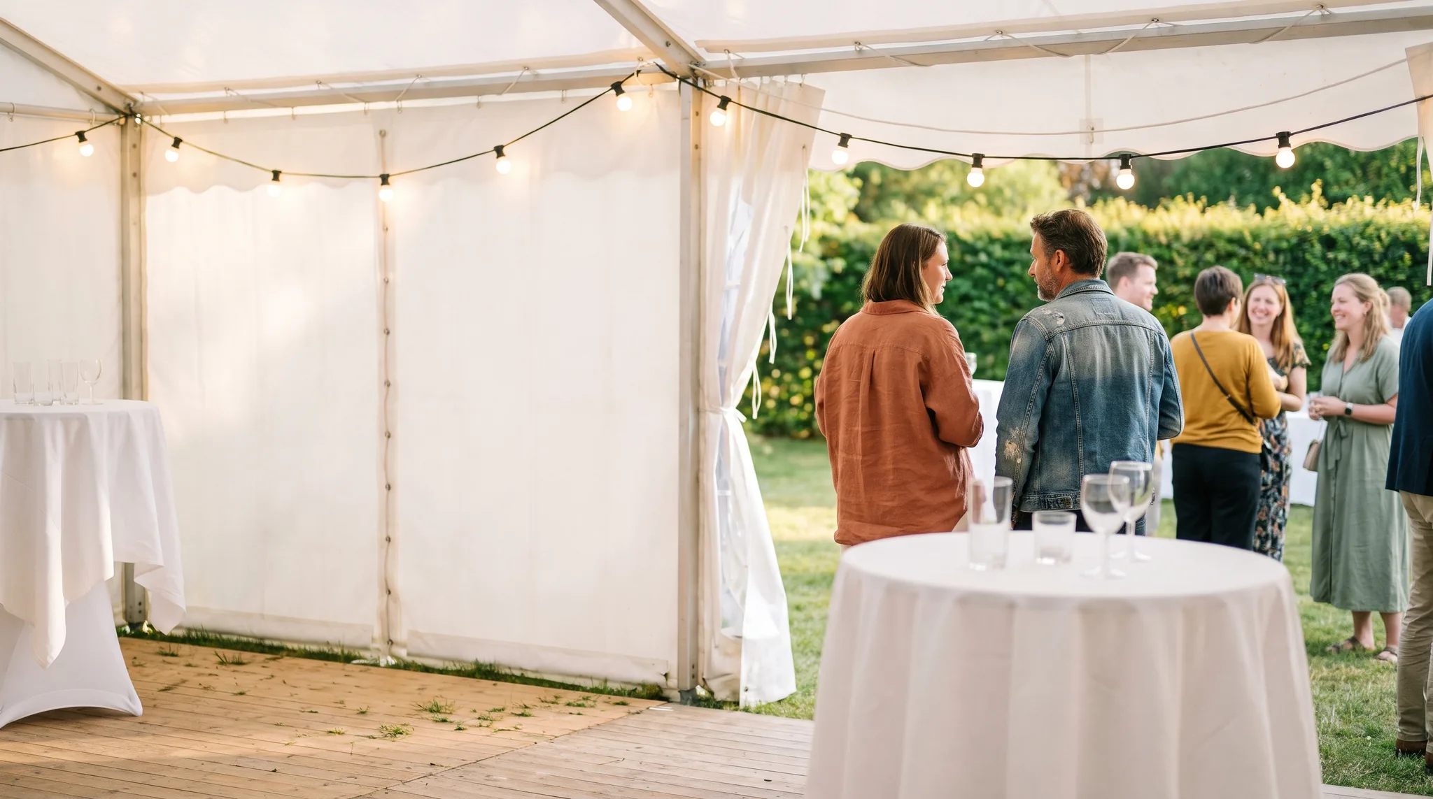 Partyzelt Detail: Holzboden unter dem Zelt mit zwei Stehtischen, Lichterkette an der Zeltstange und Blick durch die geöffnete Seitenwand in den Garten