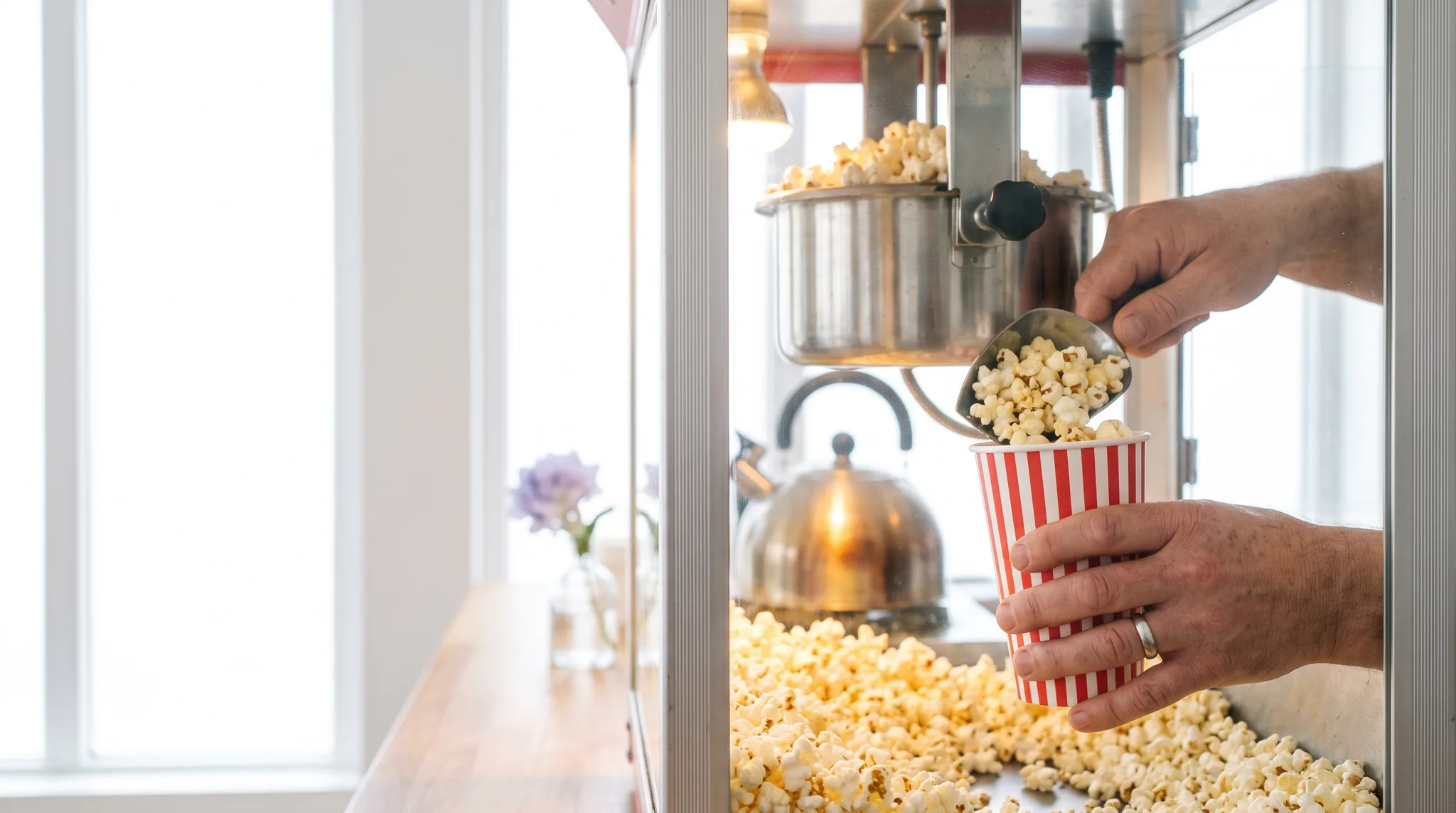 Popcornmaschine mieten Detail: Hand füllt mit einer Schaufel frisches Popcorn in einen gestreiften Becher aus der Glasvitrine der Maschine