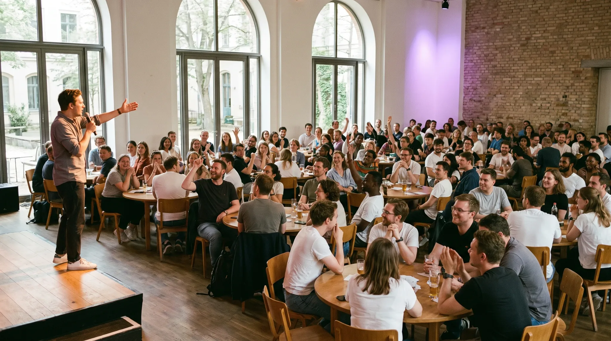 Gameshow Berlin: charismatischer Moderator mit Mikrofon vor begeisterten Teams an runden Tischen, warmes Bühnenlicht, lebhafte Stimmung.