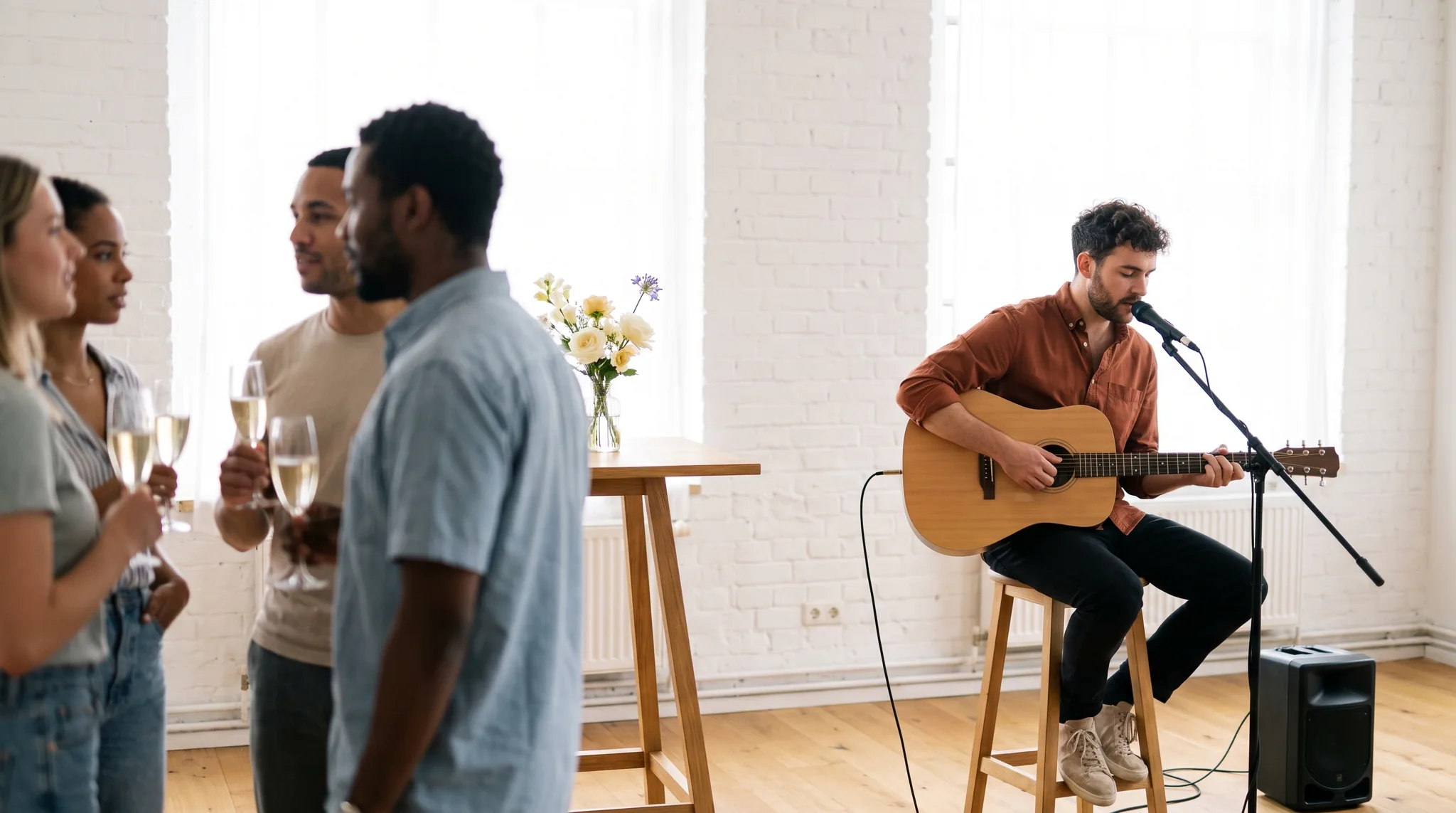 Sänger mit Akustikgitarre singt live beim Empfang einer Firmenfeier in einem hellen Loft, vorn unterhalten sich Gäste mit Sektgläsern