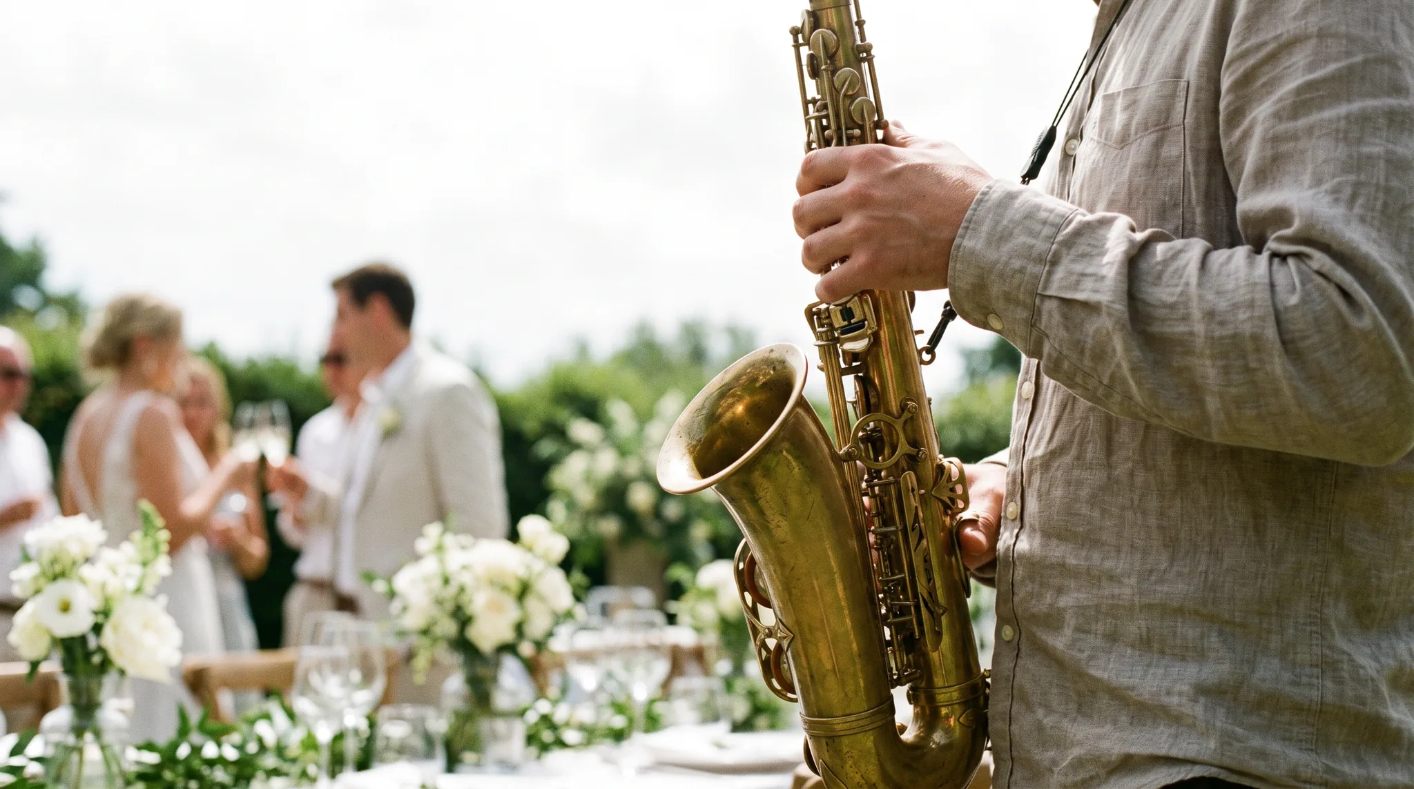 Hände eines Musikers halten ein goldenes Tenorsaxophon mit Patina im Sonnenlicht, dahinter unscharf ein Brautpaar beim Anstoßen und weiße Blumen auf der Hochzeitstafel