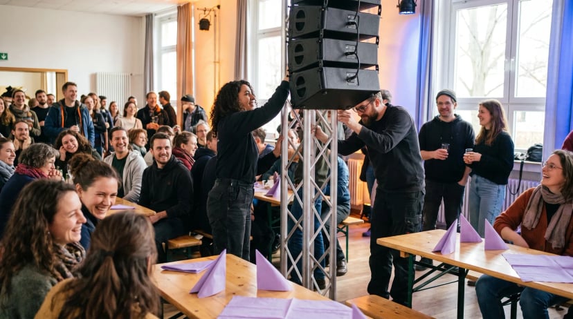 Veranstaltungstechnik Köln: Zwei Techniker hängen Lautsprecher an eine Traverse, Gäste im hellen Saal schauen zu.