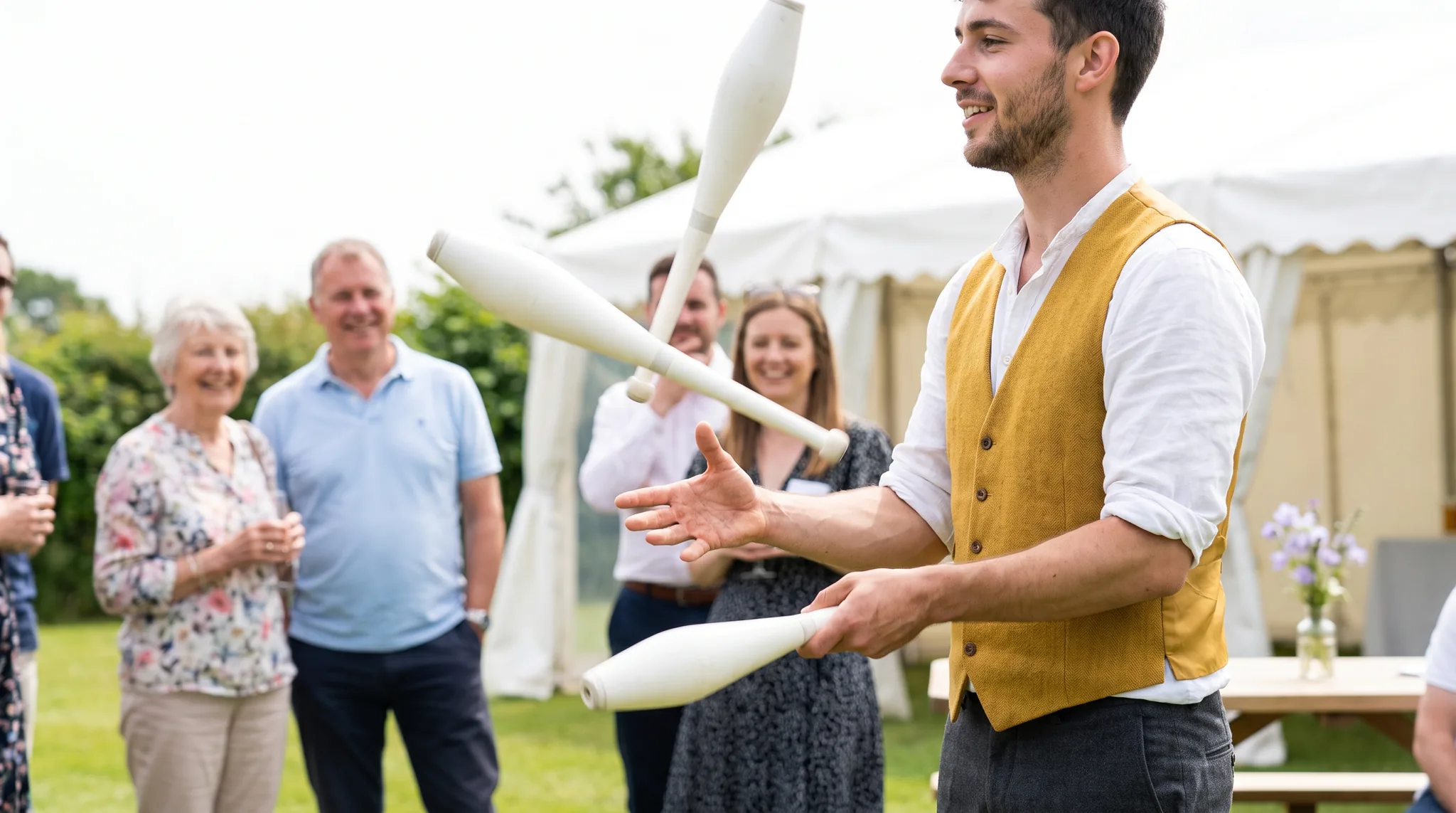 Jongleur in senfgelber Weste wirft drei weiße Keulen auf einem Sommerfest im Garten, dahinter lächelnde Gäste vor einem weißen Zelt